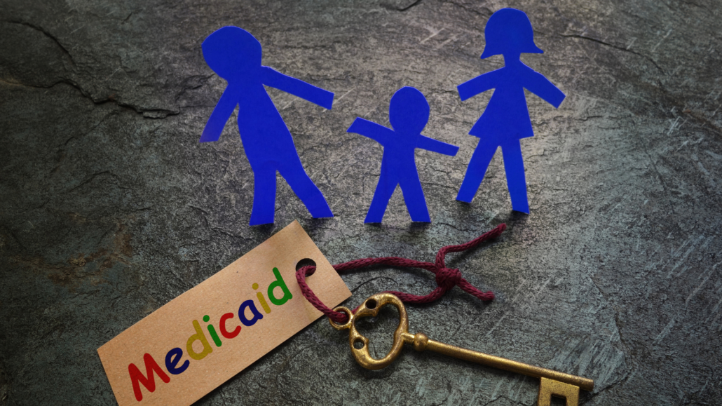 Blue paper cutout of a family on a table, accompanied by a golden key with a kraft paper label that says "Medicaid" attached by a purple yarn. 